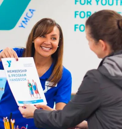 A smiling YMCA staff member in a blue polo hands a Membership & Program handbook to a woman in a black rain coat