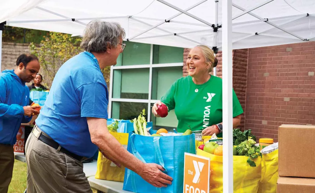 A YMCA volunteer wearing a green shirt puts an apple into a reusable grocery bag filed with produce as a man in a blue shirt and a YMCA lanyard comes to lift the bag off the table